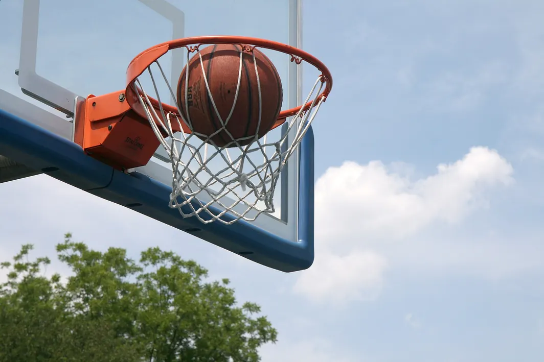Bola de basquetebol parada no aro de um cesto ao ar livre, com céu azul e nuvens