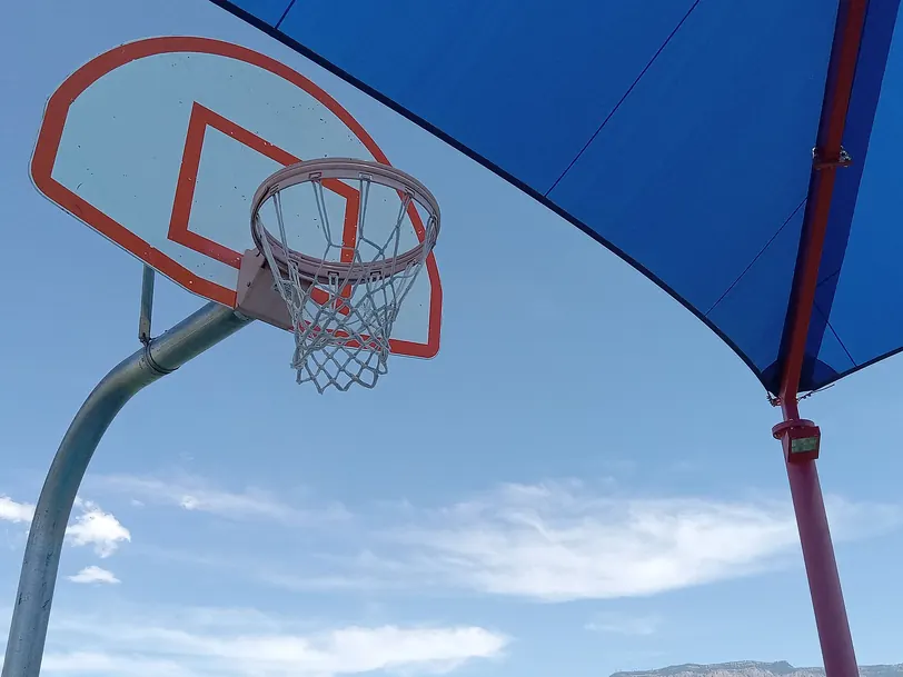 Cesto de basquetebol visto de baixo, com a tabela branca e um toldo azul a cobrir parte do céu